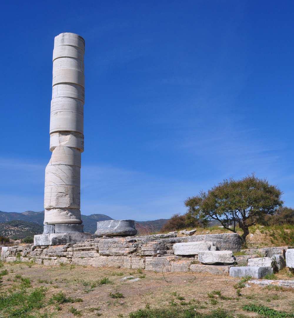 A tall, ancient stone column at the Heraion archaeological site in Pythagorion, Samos, Greece, under a blue sky.