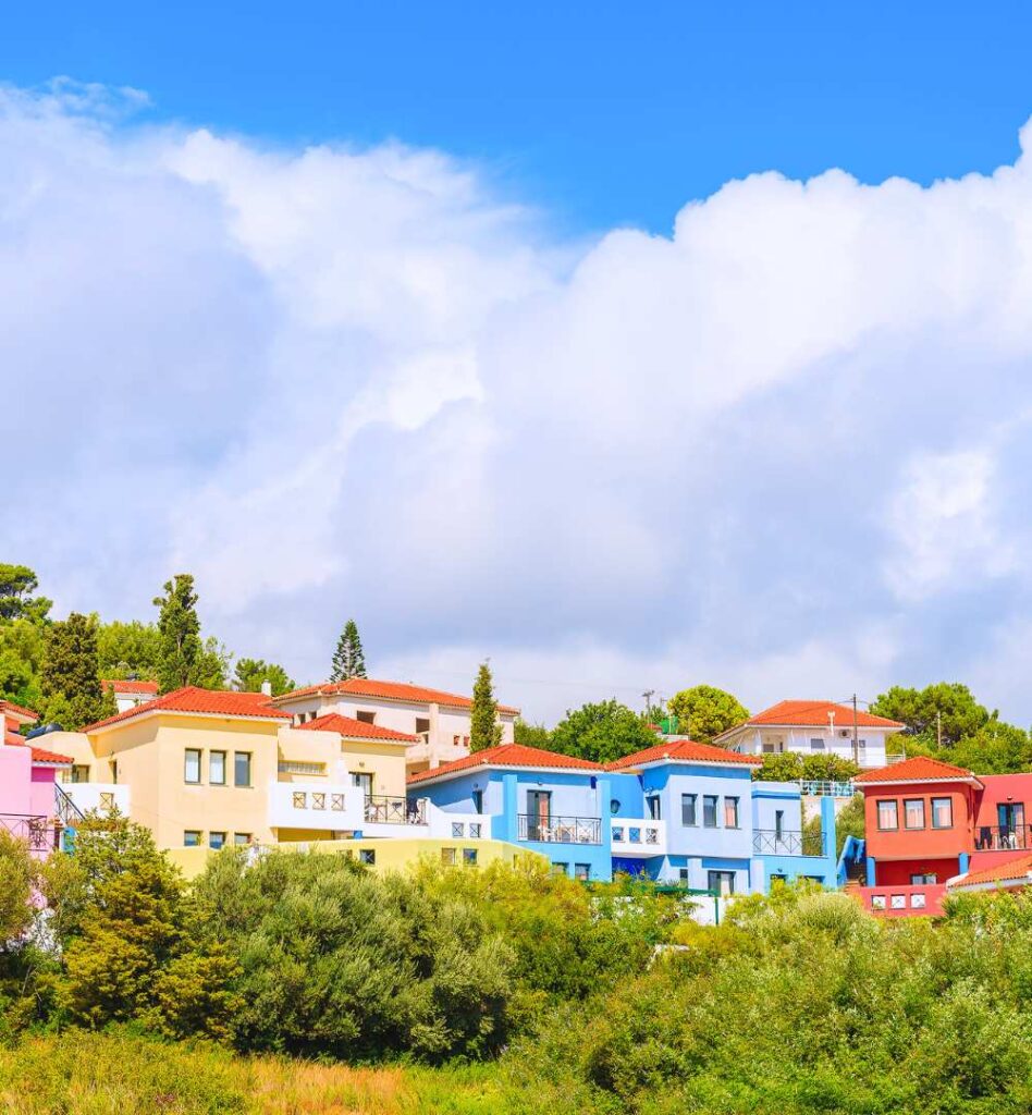 A row of colorful traditional houses in yellow, blue, and red with orange tiled roofs nestled in the greenery of Samos, Greece, under a bright sky.