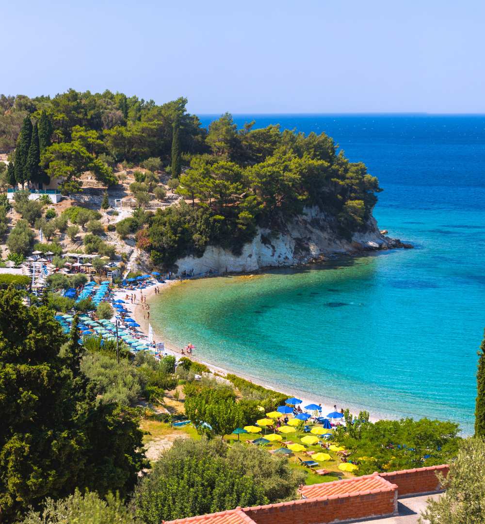 Lush green hillside curving around the pebble shores of Tsamadou Beach, Samos, Greece, with sun umbrellas.