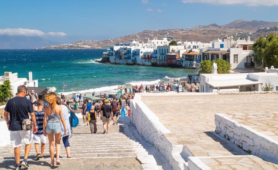 Many people walking down the whitewashed steps  with view in the sea.
In the background colorful houses in Little Venice Mykonos Island.