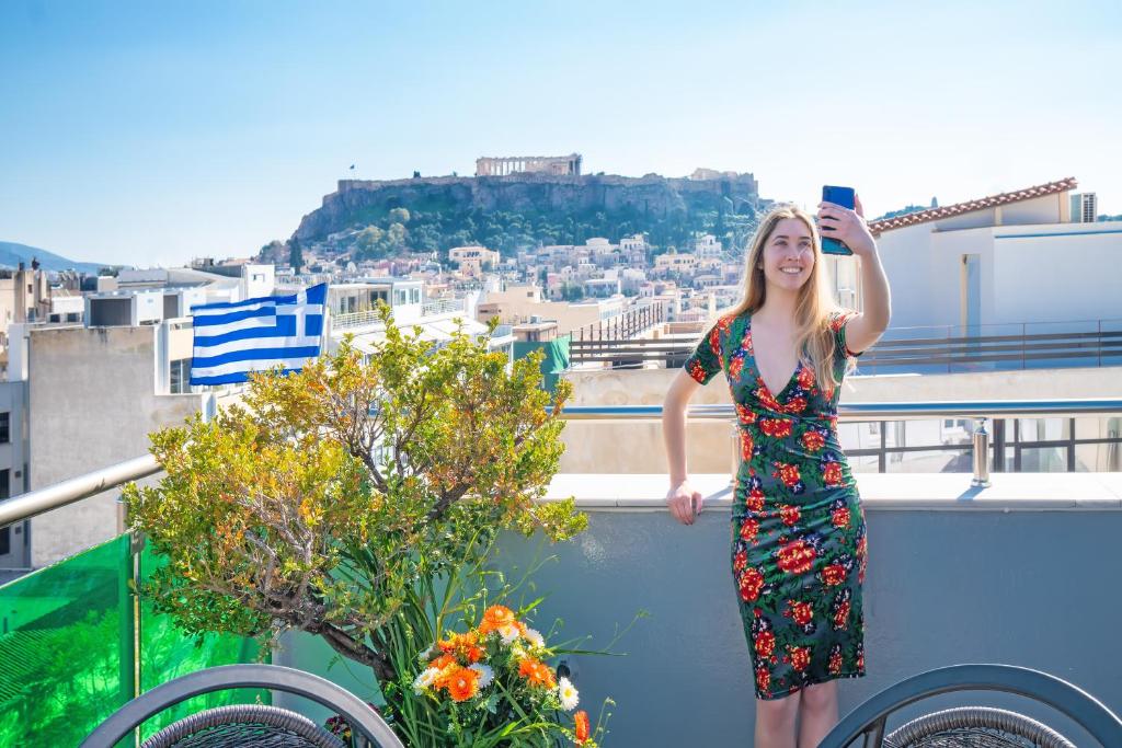 A blond woman in a balcony taken a selfie, the Greek Flag and a view to Acropolis in Athens.