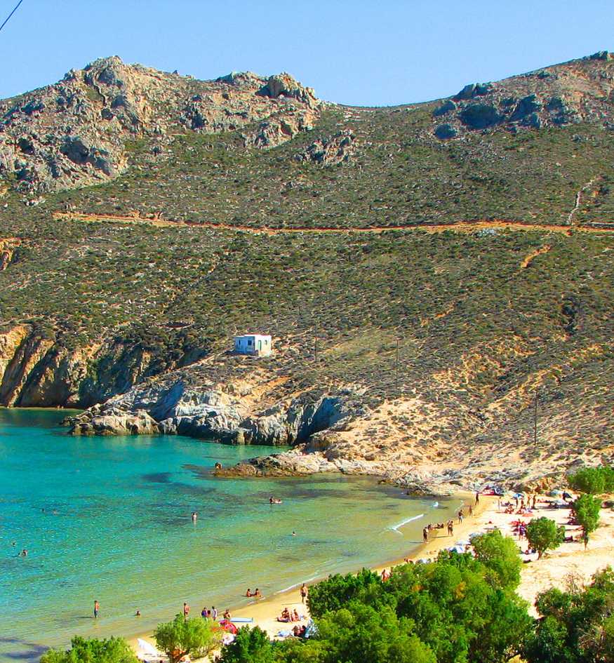 People relaxing on a sandy beach with turquoise water and green tamarisk trees under rugged hills in Serifos, Greece.