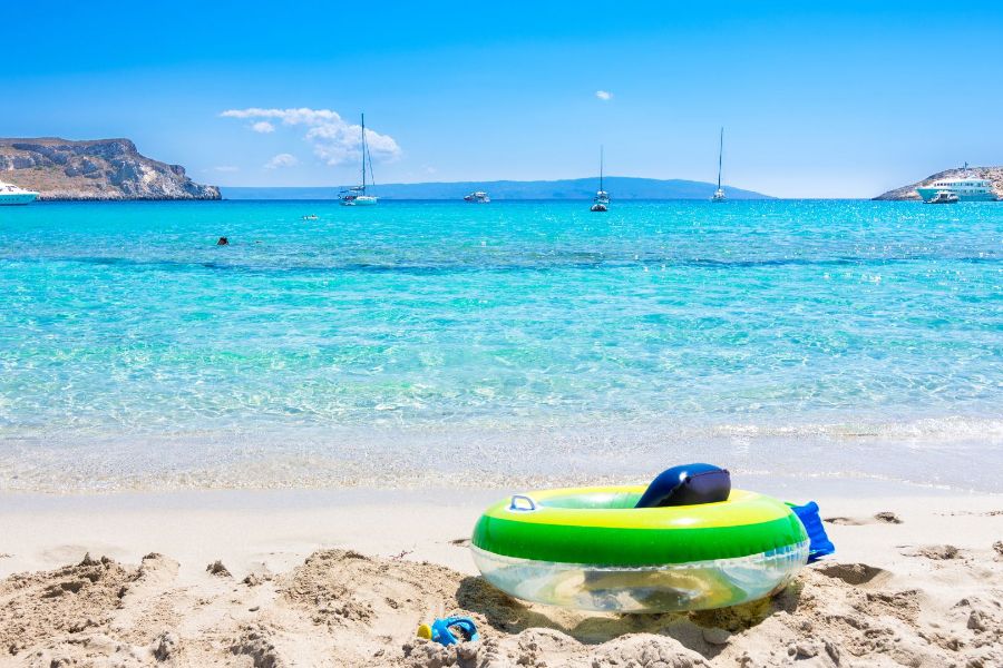 A sandy beach on Elafonisos island in Greece with turquoise water, sailboats in the distance, and an inflatable ring on the shore.
