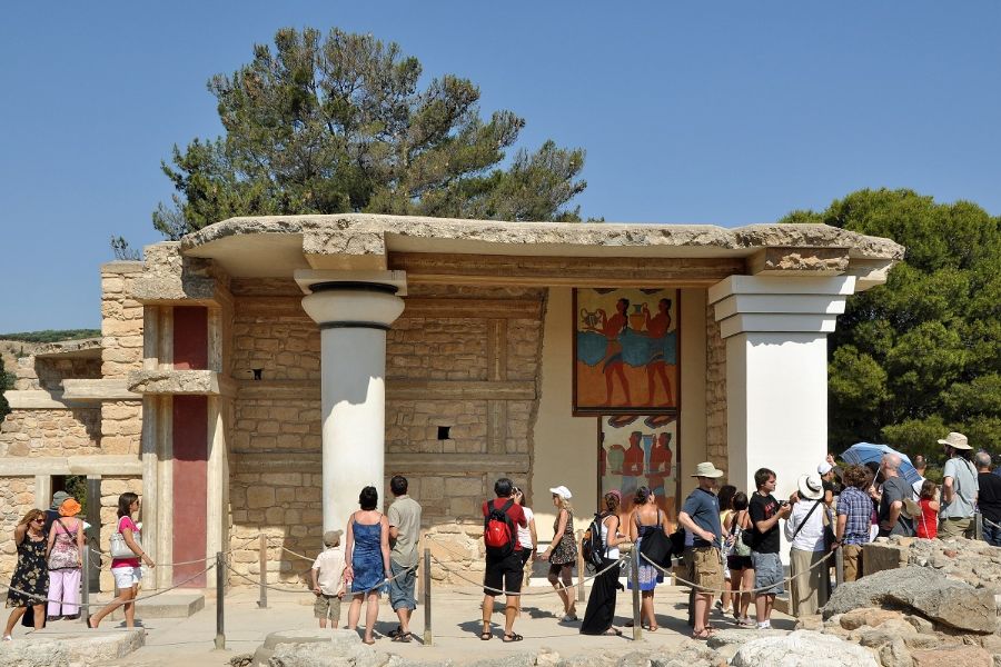 Visitors exploring the ancient ruins of Knossos Palace in Crete, Greece, with its iconic columns, frescoes, and historical architecture under a clear blue sky.