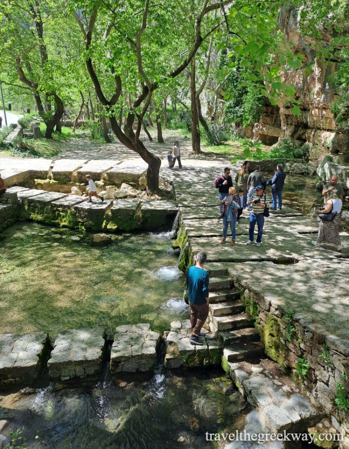 Crowds gathered at Xenia Cafe by the river, surrounded by tall plane trees and stone buildings in Livadia.