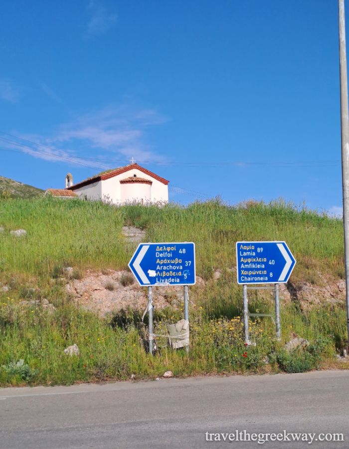 Road signs pointing to Livadia, Delphi, and Arachova with a small church on a green hill under a clear blue sky.