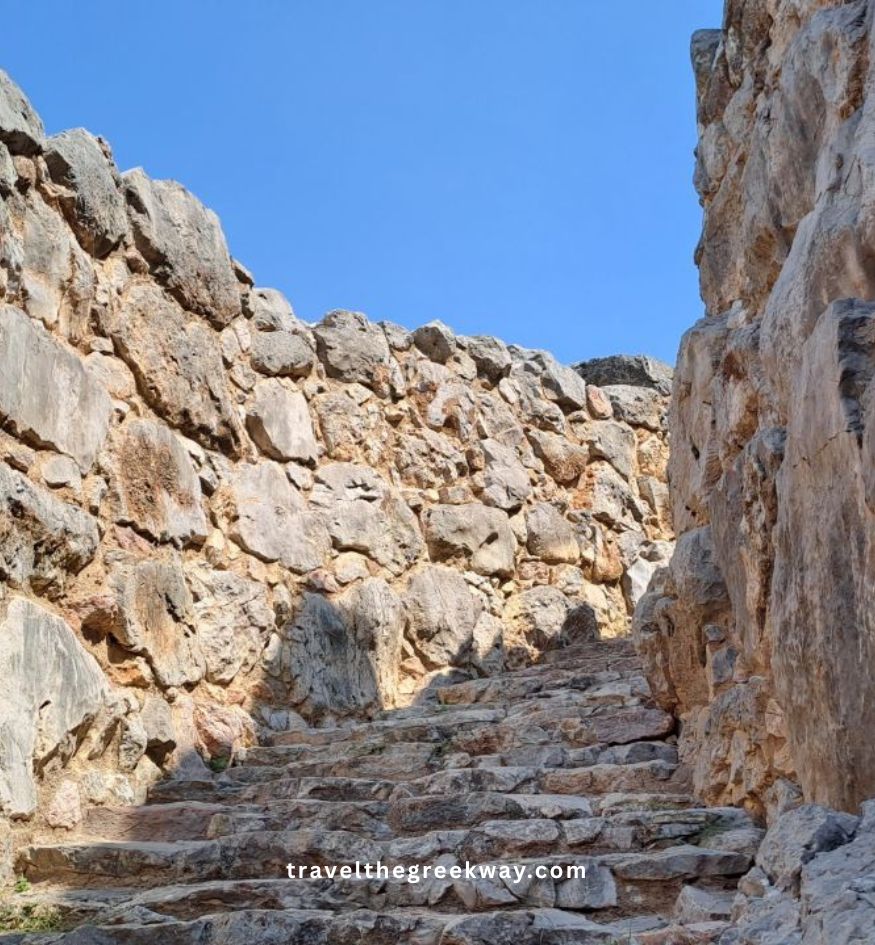A low-angle shot looking up a steep stone staircase framed by massive, ancient limestone walls at the Citadel of Tiryns, near Nafplio, Greece.