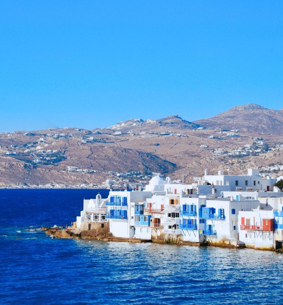 Little Venice in Mykonos with white Cycladic houses, colorful balconies, turquoise water, and hills in the background.