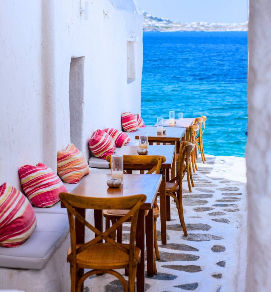 Seaside café in Mykonos with wooden tables, colorful cushions, white walls, and bright blue Aegean Sea in the Cyclades, Greece.