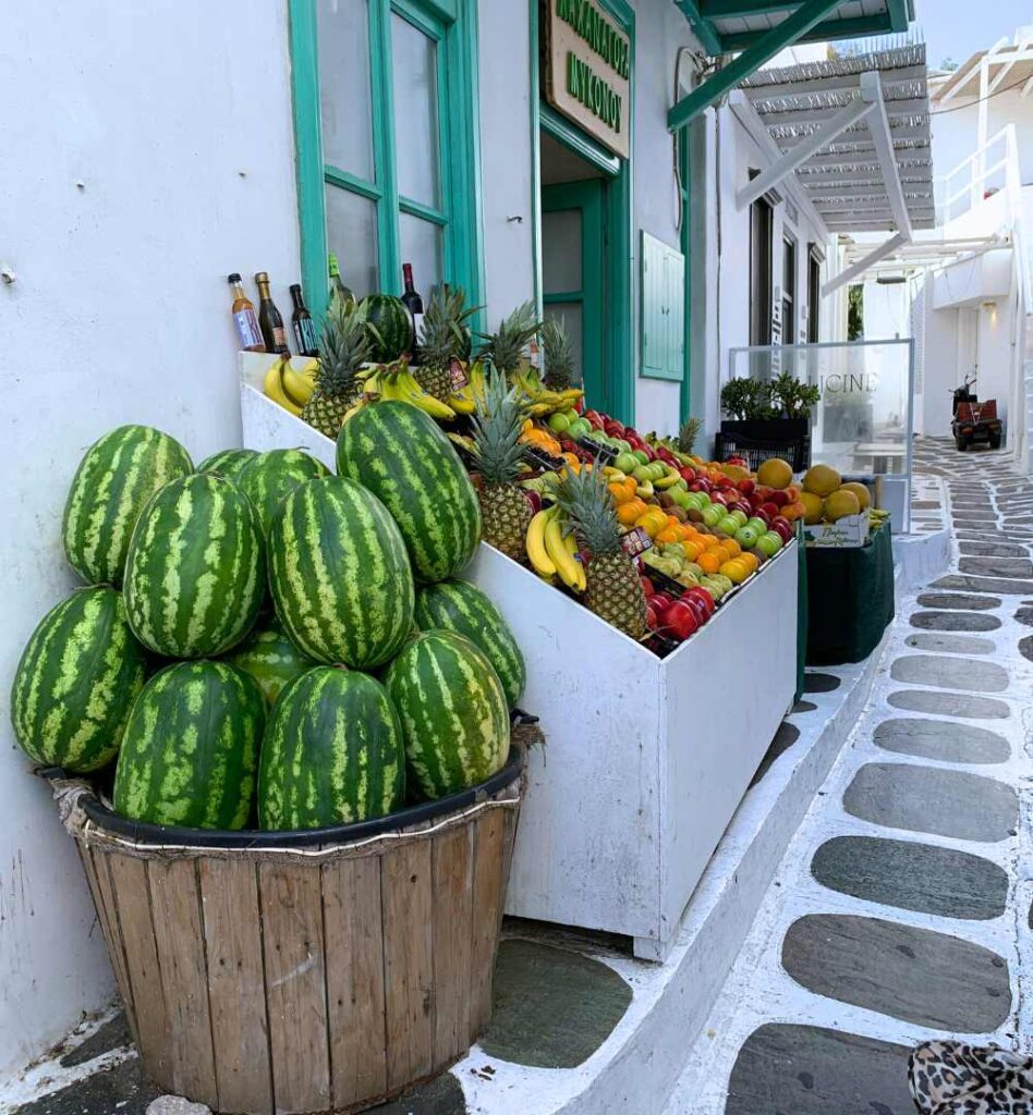 Traditional Greek grocery shop in Mykonos with fresh fruit displayed along a whitewashed street.