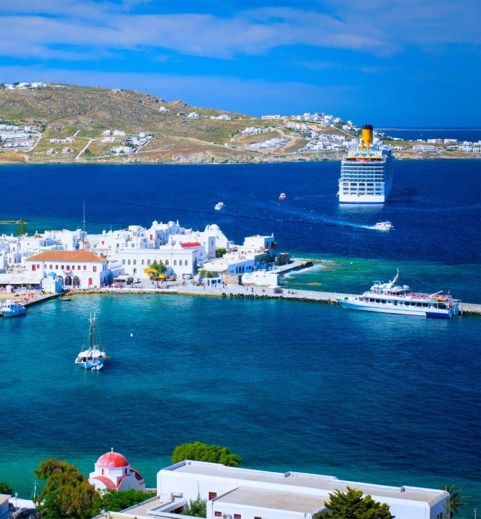 Mykonos Old Port with whitewashed buildings and cruise ships anchored offshore.