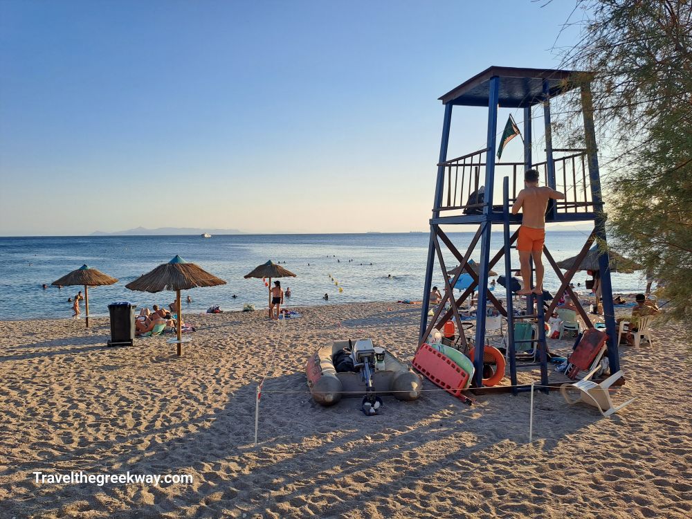 A lifeguard stands on a wooden tower overlooking a sandy beach with straw umbrellas and people swimming in the Paleo Faliro sea.