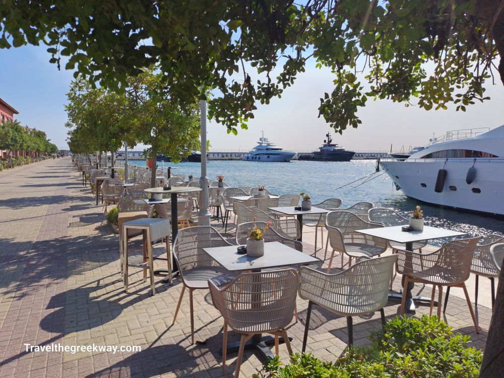 A waterfront promenade at Flisvos Marina, lined with outdoor cafe seating, overlooking docked yachts and the sea.