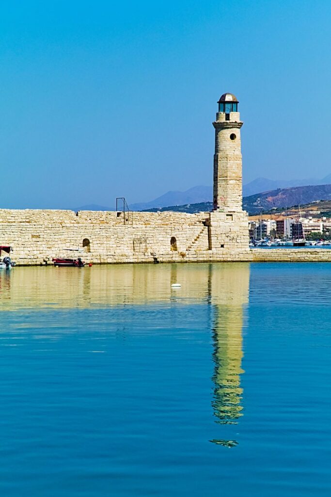 Stone lighthouse at the end of the pier in Rethymno, Crete, reflected in the calm, blue waters of the harbor with distant mountains in the background.