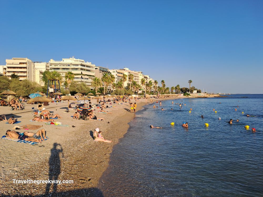 Beachgoers at Paleo Faliro Beach, Athens Riviera, with swimmers in the sea and modern buildings in the background.