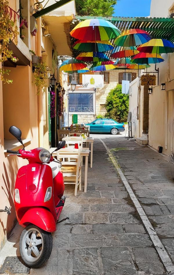 Colorful narrow street in Rethymno, Crete, Greece, featuring a red scooter parked by a wall, tables set for dining, and vibrant umbrellas hanging overhead