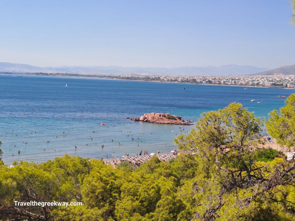 Athens Riviera beach with sunbathers, umbrellas, and a rocky islet in the clear blue water.