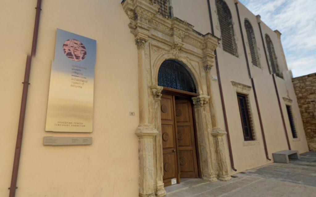 Entrance to the Archaeological Museum of Rethymno, Crete, featuring a grand wooden door framed by ornate stonework and a metal plaque displaying the museum's name in Greek and English.