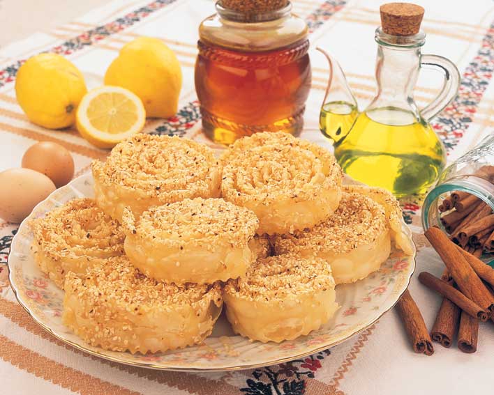 Traditional Cretan dessert, Xerotigana, arranged on a plate with ingredients like honey, olive oil, lemons, cinnamon sticks, and eggs displayed around.