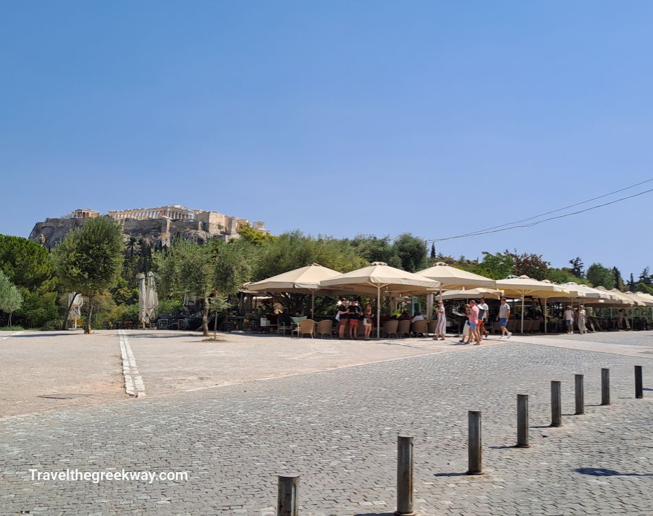 A view of the Acropolis from Apostolou Pavlou Street, with outdoor seating shaded by umbrellas and trees lining the street.