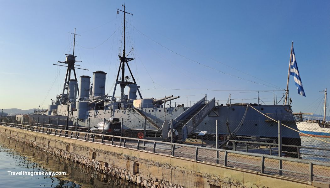 The historic Averof battleship docked at Flisvos Marina, with the Greek flag flying at the stern, under clear blue skies.