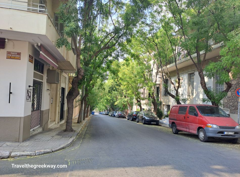 A tree-lined, quiet residential street in Athens, Greece, with parked cars and shaded sidewalks.