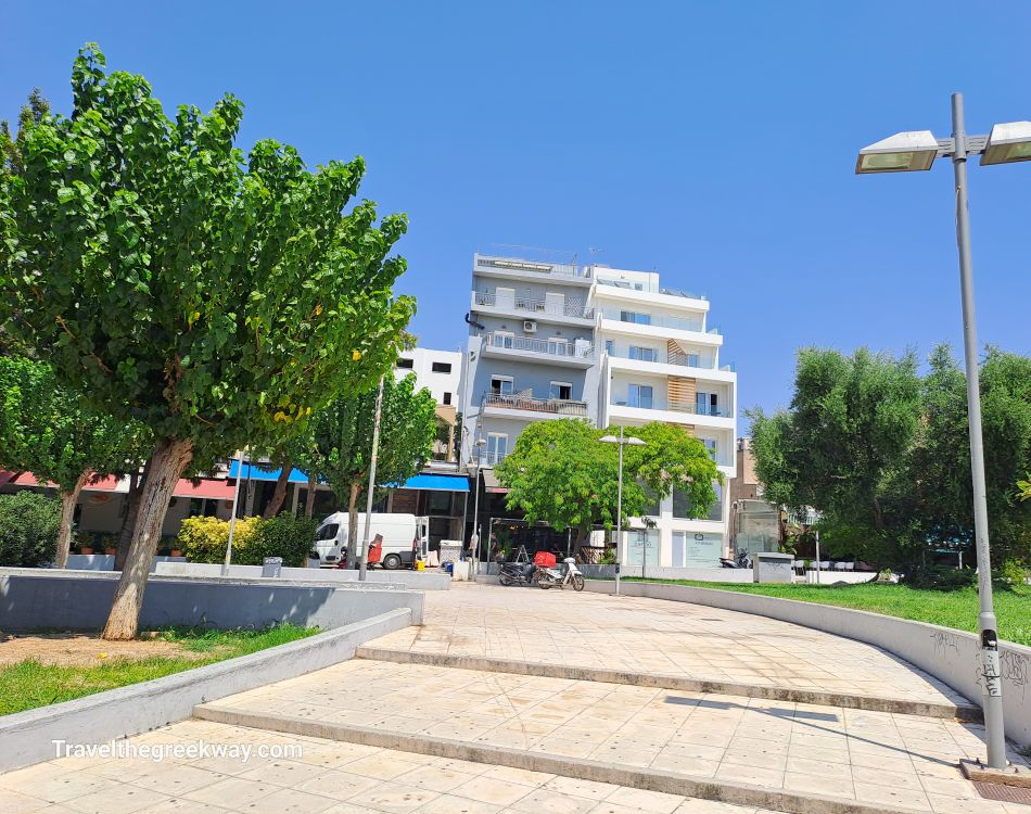 Open plaza area with green trees and a white modern building in the background in Athens, Greece.