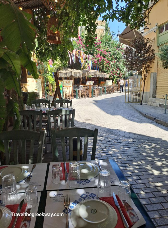  A charming outdoor dining setup at a tavern in Psiri, Athens, with tables set for lunch under hanging plants and colorful decorations.