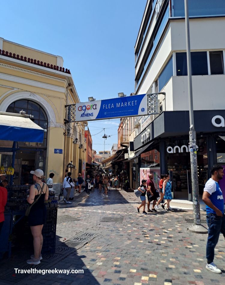 The bustling entrance to Monastiraki Flea Market in Athens, with people walking under a sign and into the market's busy streets.