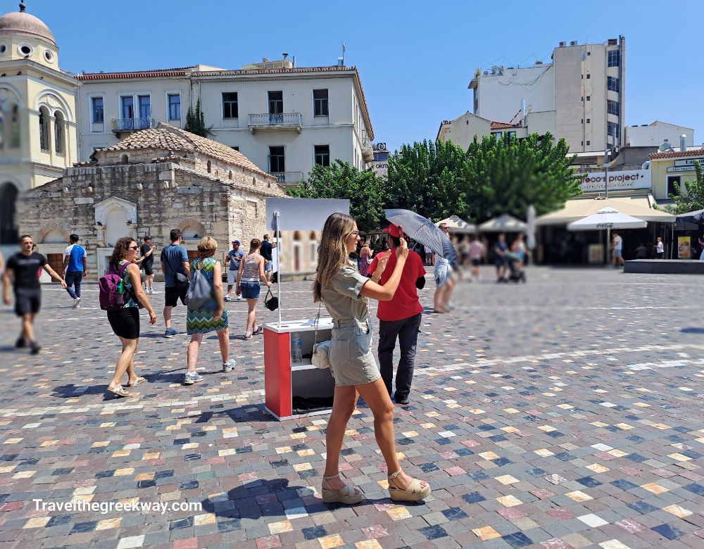 A busy scene in Monastiraki Square, Athens, with tourists and locals walking near historic buildings and a small church under a bright sky.