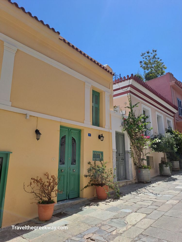 Charming neoclassical houses in Plaka, Athens, painted in soft pastel colors with green doors and potted plants lining the street.