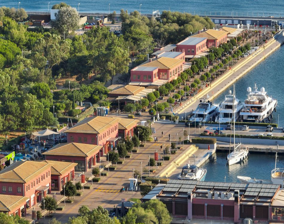 An aerial shot of Flisvos Marina showcasing the neatly arranged red-roofed buildings, yachts docked in the marina, and lush green surroundings.