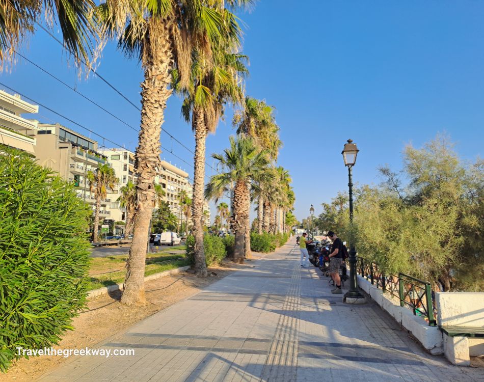 Palm-lined promenade at Flisvos Marina, Athens Riviera, with people walking and modern buildings nearby.