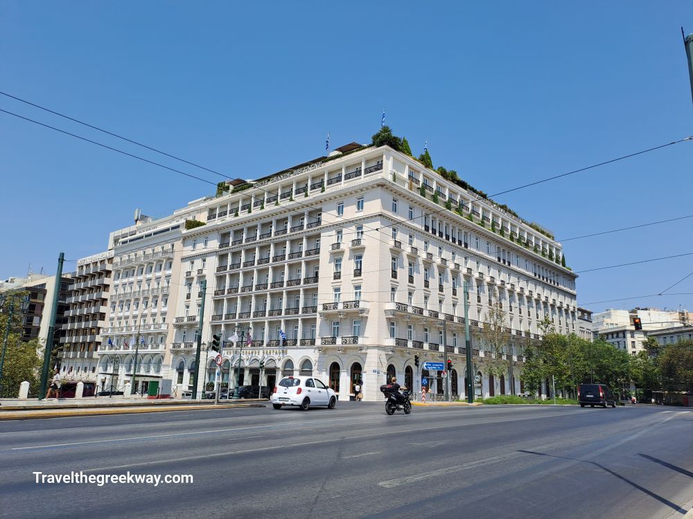 The historic Grande Bretagne Hotel in Syntagma, Athens, with its elegant neoclassical architecture and a bright blue sky overhead.