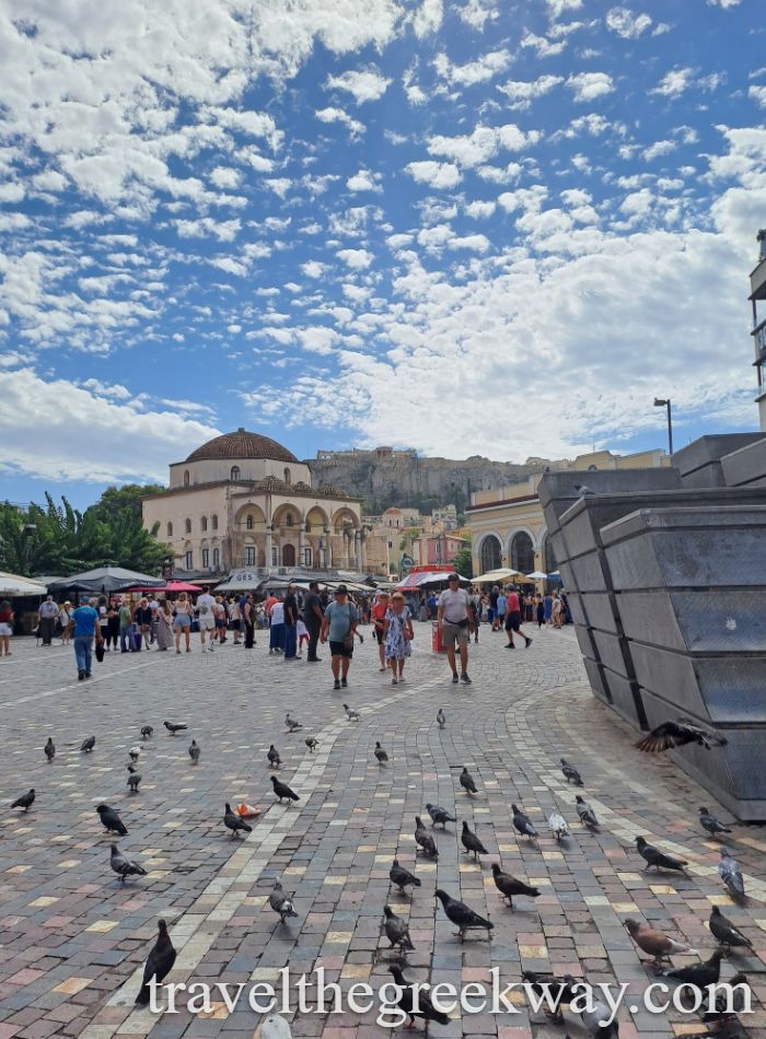 Busy Monastiraki Square in Athens with pigeons on cobblestone streets, tourists walking, and the Acropolis in the background.