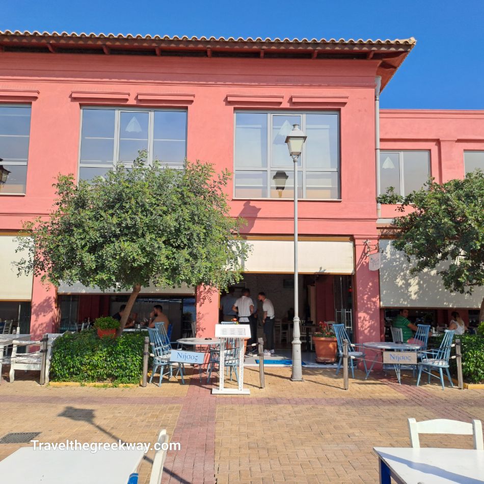  The exterior of Nisos restaurant in Flisvos Marina, featuring a rustic red building with outdoor seating under the shade of trees.