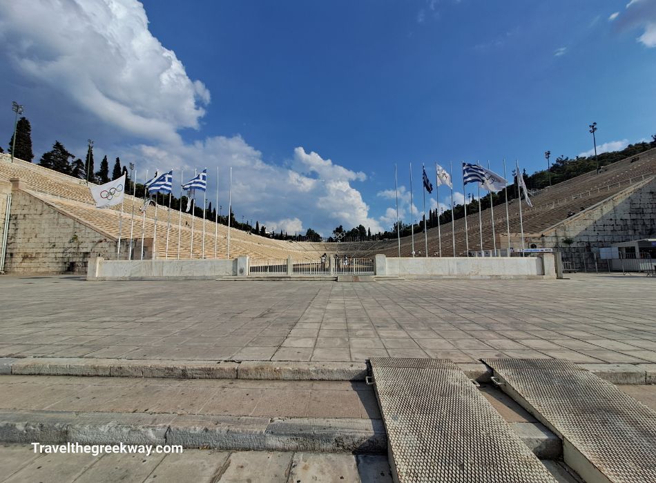 The historic Panathenaic Stadium in Athens, with Greek flags waving and a clear blue sky above.