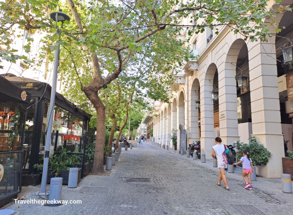 A charming, tree-lined street in Syntagma with people strolling and relaxing at outdoor cafés under the shade of green trees.