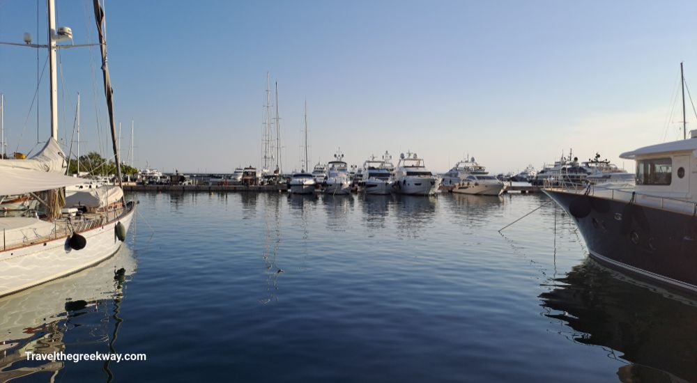 A view of several luxurious yachts docked in Flisvos Marina, with calm waters reflecting the vessels and a clear sky overhead.