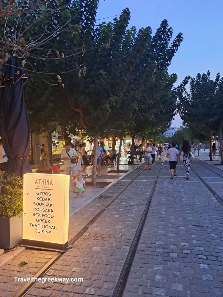 Evening scene in Athens with people walking along a cobblestone street lined with trees and illuminated restaurant signs.