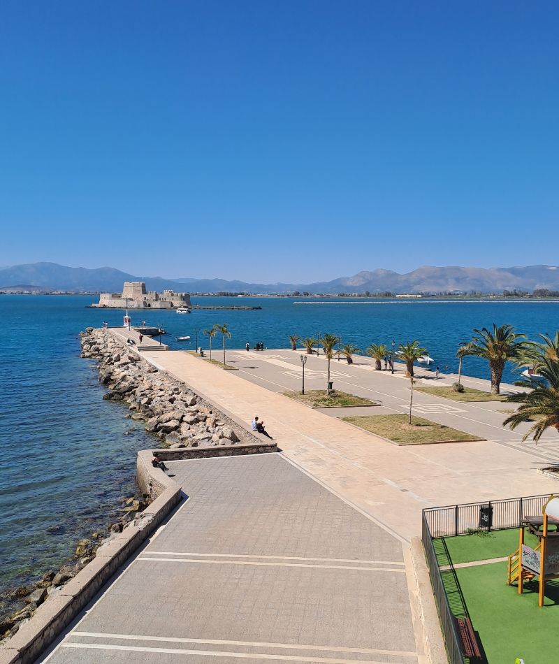 Nafplio’s seafront promenade leading to Bourtzi Castle with mountains and the blue sea in the background.