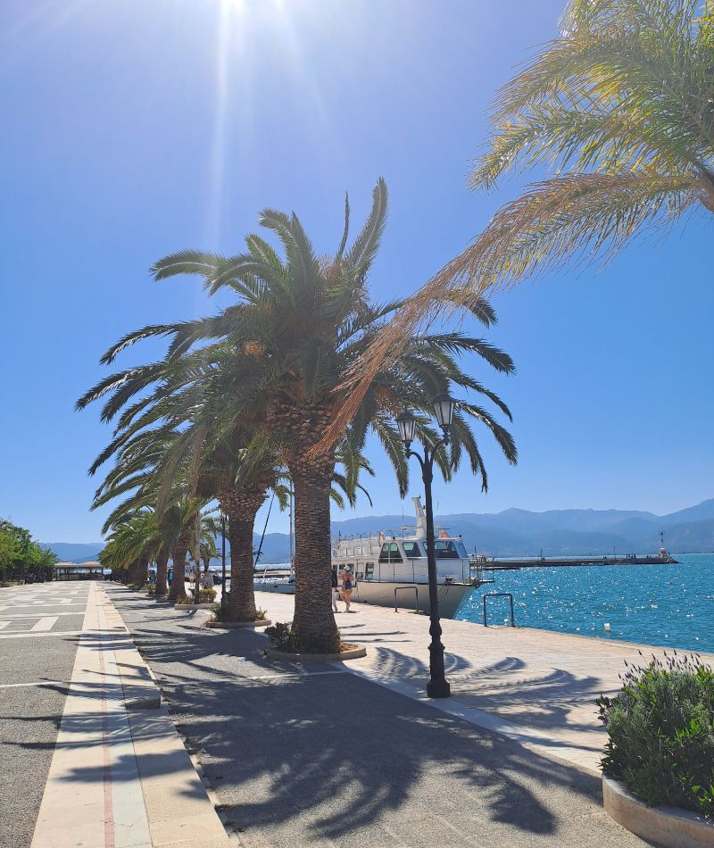 Sunny promenade in Nafplio lined with tall palm trees and a docked boat against the blue sea.