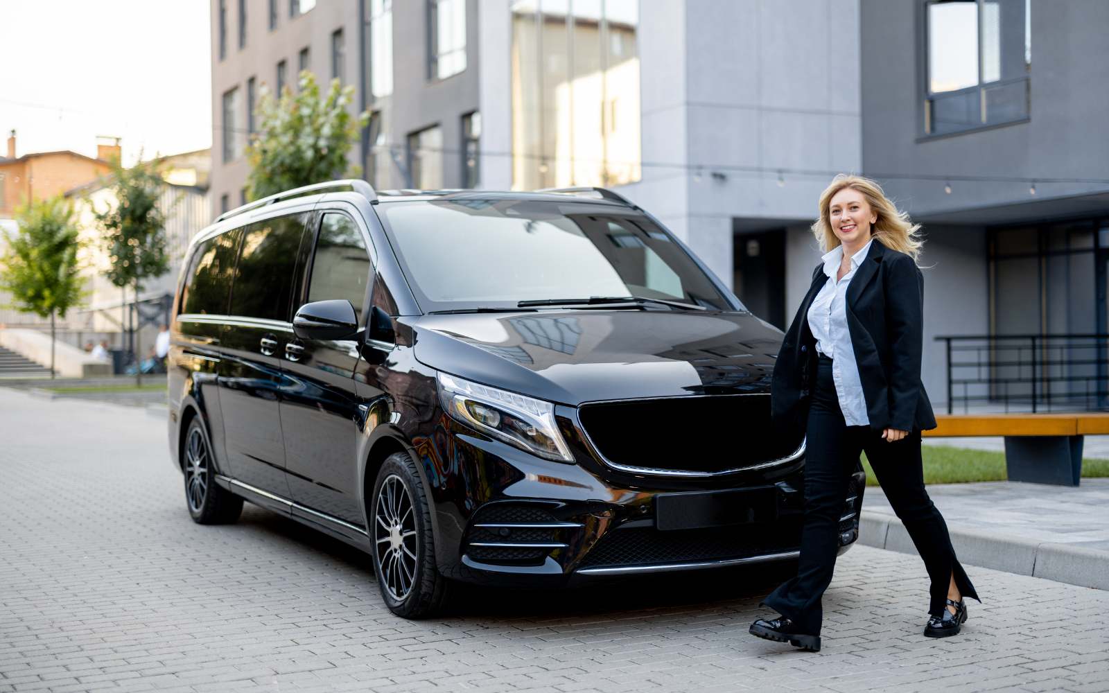 Female driver standing beside a sleek black minivan, ready for a city transfer in Greece.