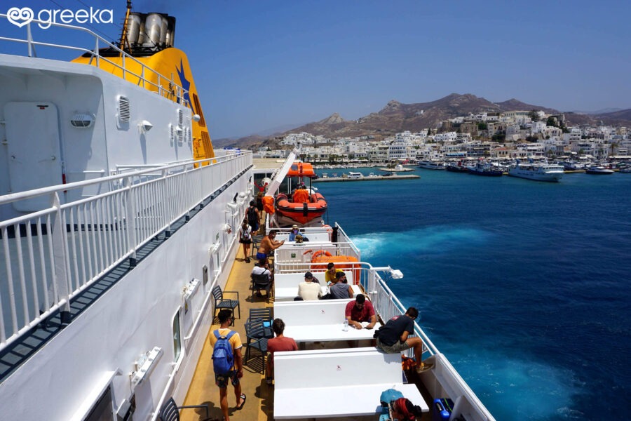 Passengers enjoying the view from the upper deck of a Blue Star Ferries ship as it approaches a Greek island port with white houses and hills.