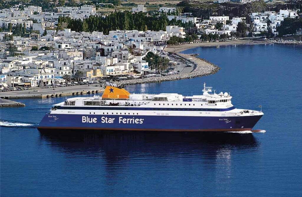 Blue Star Ferries vessel sailing into a Greek island port, with whitewashed buildings and scenic coastline in the background.