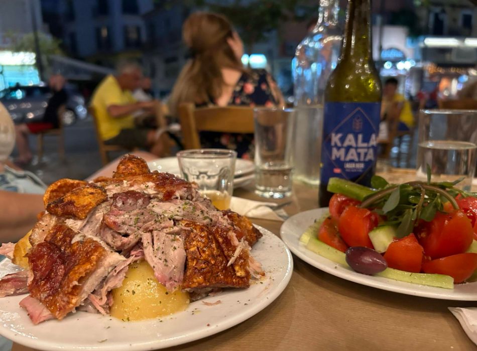 Greek roast pork dish paired with a fresh salad and Kalamata beer, enjoyed outdoors at a local restaurant.