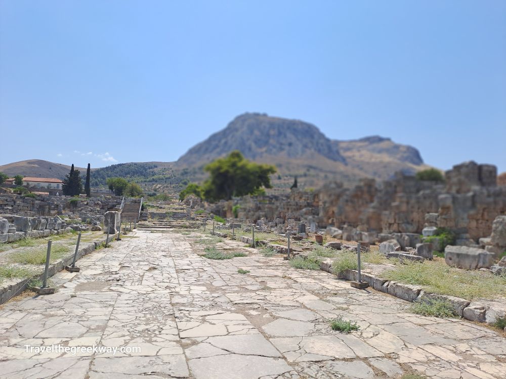  Stone-paved Lechaion Road lined with ruins and mountain landscape in the background at Ancient Corinth.