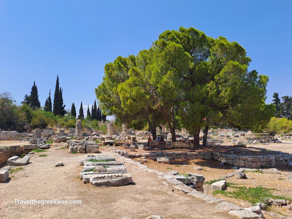 Shaded seating area under large trees amidst ruins at Ancient Corinth.