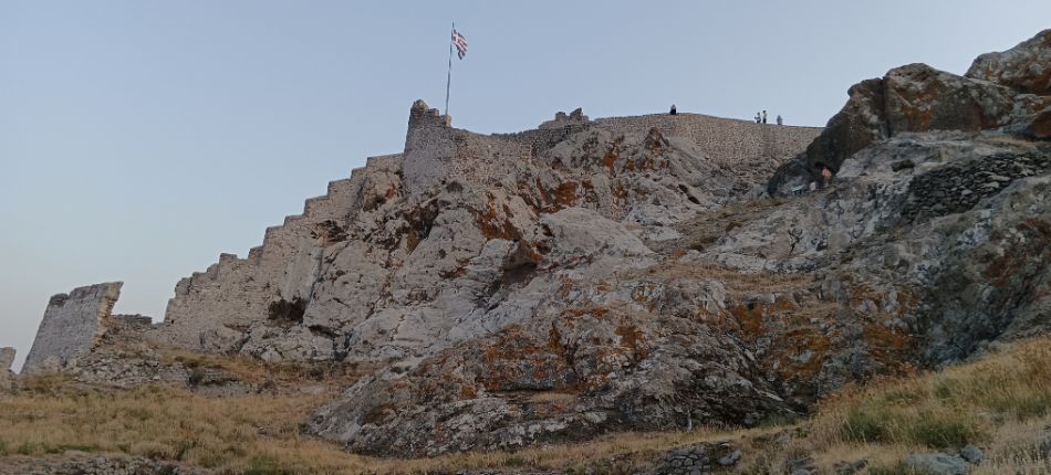 The towering walls of Myrina’s castle, with the Greek flag waving at the top, against a rocky hillside.