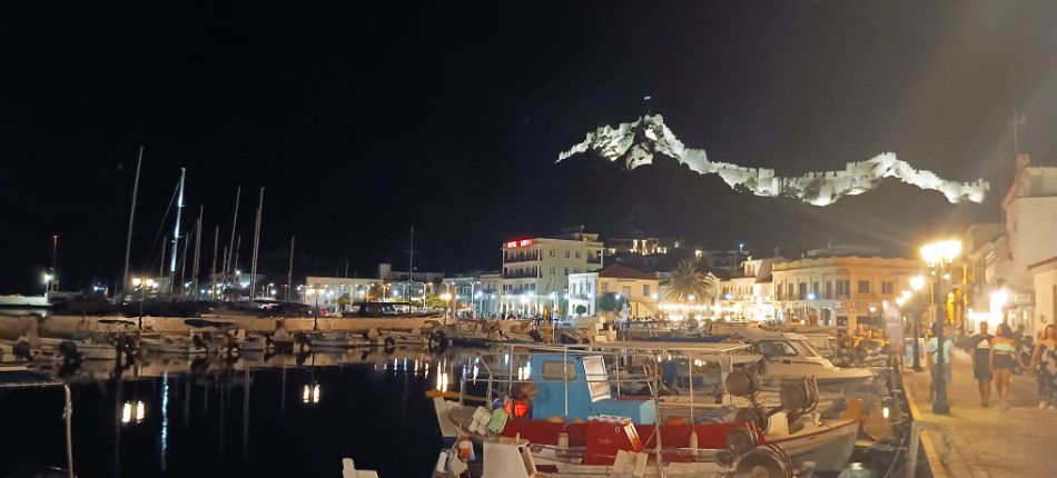 Night view of Myrina’s harbor with boats docked and the illuminated Venetian castle on the hill in the background.